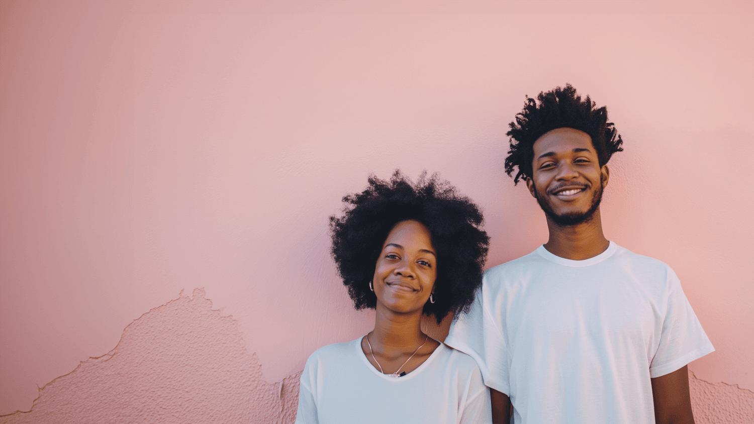 A smiling couple standing close together against a soft pink wall